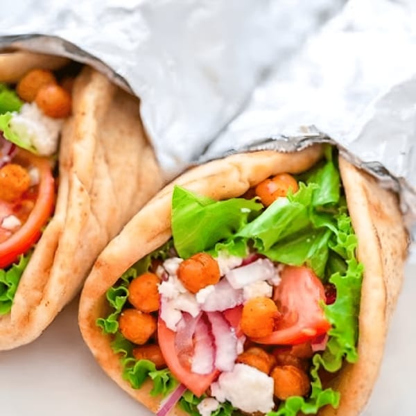 Two Greek gyros, wrapped in foil, filled with lettuce, tomatoes, feta, and crispy chickpeas.