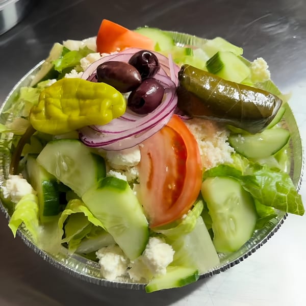 A vibrant Greek salad in a foil container, featuring lettuce, cucumbers, tomatoes, olives, feta, and a stuffed grape leaf.